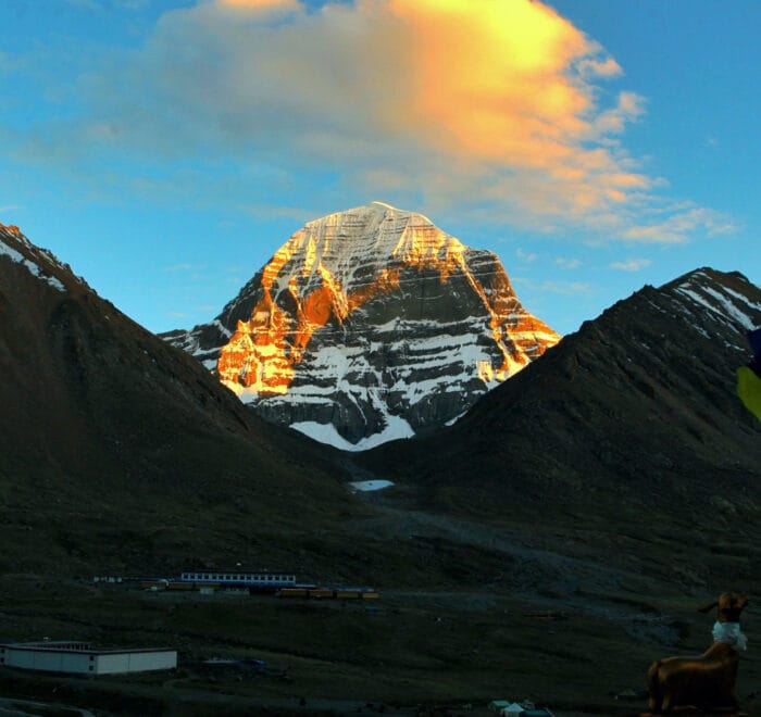 View of Mount Kailash at sunrise, as seen from Drirra Put Monastery, with the first light illuminating the snow-capped peak and casting a warm glow over the surrounding mountains and landscape.