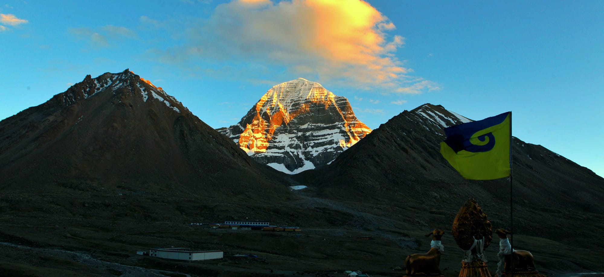 View of Mount Kailash at sunrise, as seen from Drirra Put Monastery, with the first light illuminating the snow-capped peak and casting a warm glow over the surrounding mountains and landscape.