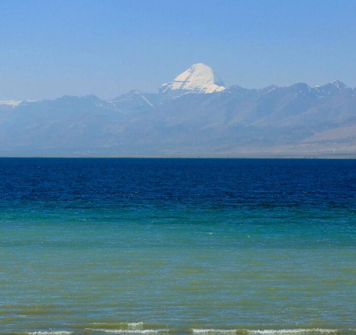 A panoramic view of Mount Kailash and Lake Mansarovar, with the snow-capped peak of Kailash towering over the tranquil, clear blue waters of the lake, surrounded by rugged mountains and open skies, capturing the majesty and sacredness of this revered site.